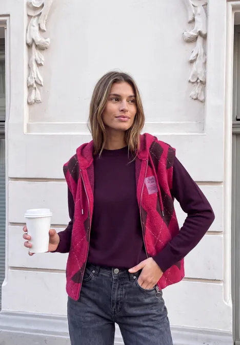 Frau vor hellem Wandrelief, trägt rote Weste, hält Kaffeebecher, lässig in Jeans und Pullover.