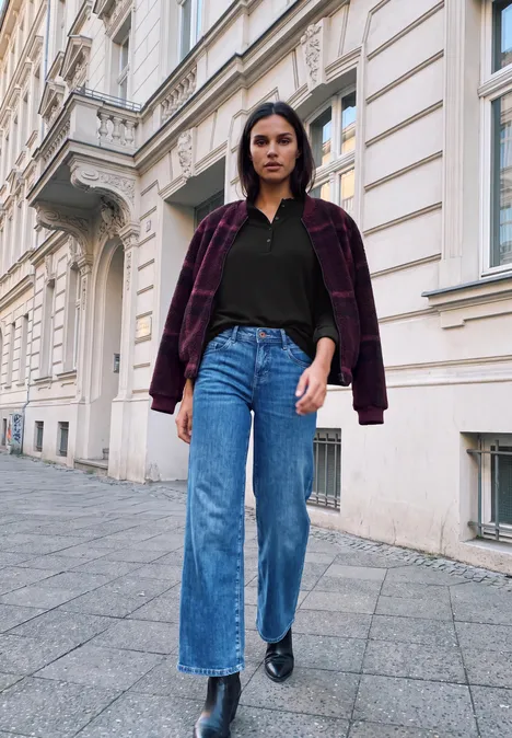 Eine Frau in weiten Jeans, schwarzem Shirt und karierter Jacke geht an einer Altbau-Fassade vorbei.