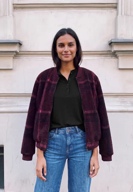 Eine Frau in Jeans, schwarzem Shirt und weinrotem Karomantel steht vor einer hellen Hauswand.