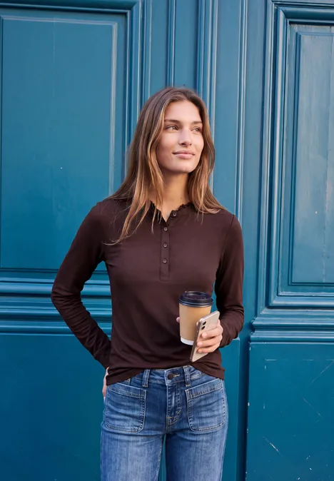 Langarm Shirt mit Rundhals und Rüschen russet red Junge Frau lehnt vor türkisem Türflügel, trägt braunes Longsleeve, Jeans und hält Kaffeebecher, lächelt.