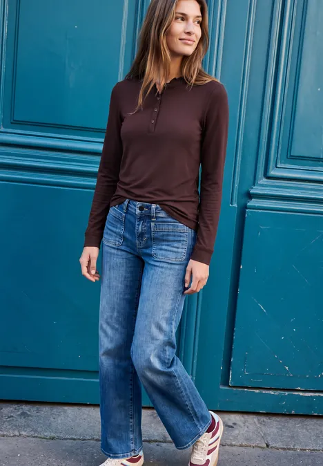 Langarm Shirt mit Rundhals und Rüschen russet red Frau steht vor einer blauen Tür, trägt braunes Oberteil, Jeans und Sneaker, lächelt und schaut zur Seite.