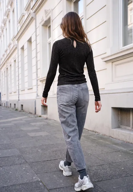 Eine Frau in schwarzem Pullover und grauer Jeans spaziert an einer weißen Häuserfassade entlang.