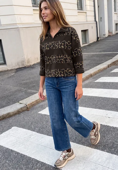 Femme en chemisier à motif, jean droit et baskets traverse un passage piéton en ville.