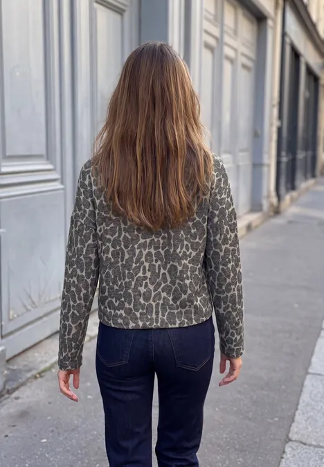 Frau mit langen braunen Haaren, gemusterter Jacke und Jeans geht auf Gehweg neben Häuserfront