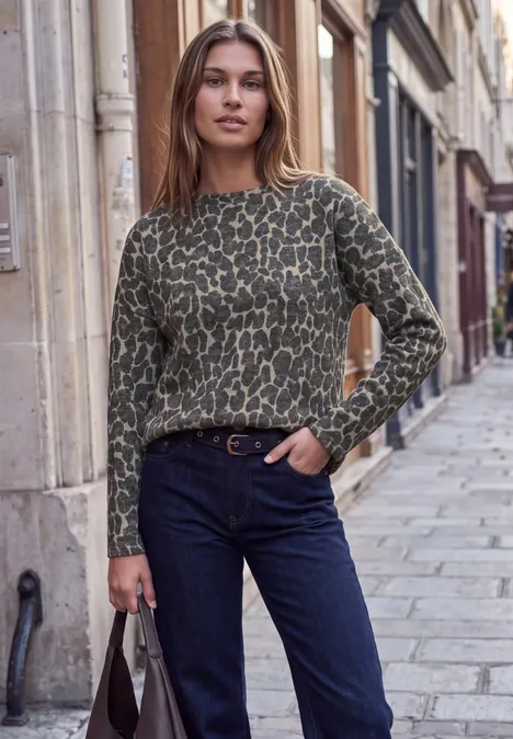 Eine junge Frau in grünem Musterpullover und Jeans posiert mit Tasche auf einer gepflasterten Straßengasse.