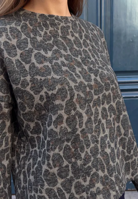 Oberkörper einer Frau in einem grauen Pullover mit Leopardenmuster vor dunklem Hintergrund.