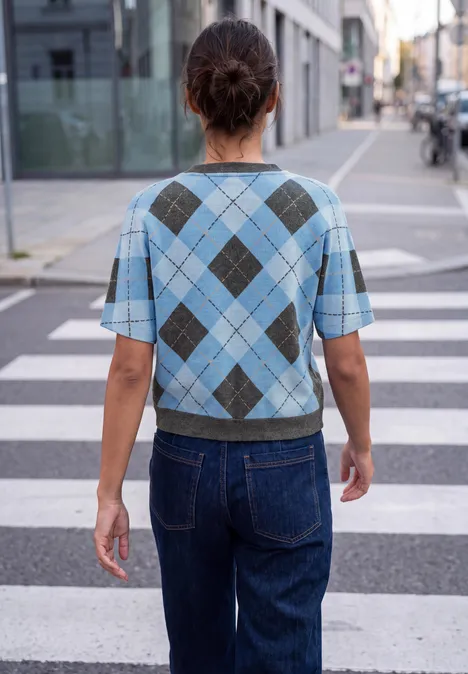 Frau in blauem Rautenmuster-Pullover und Jeans steht auf einem Zebrastreifen in der Stadt.