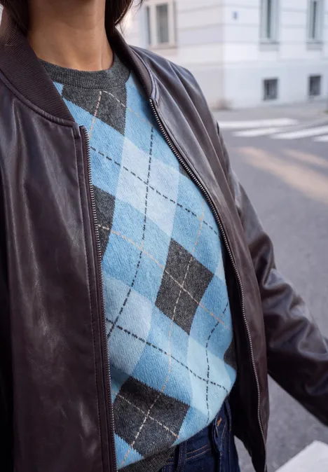 Im Bild ist eine Person mit blau-grauem Rautenmuster-Pullover und brauner Lederjacke auf der Straße.