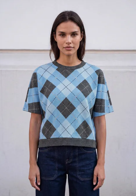 Frau trägt kurzes blau-graues Rauten-Pullover-Shirt und dunkle Jeans vor neutralem Hintergrund.