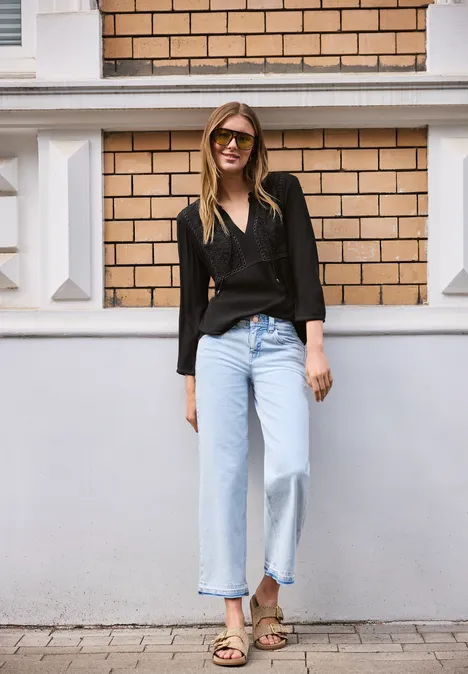 Vrouw voor een bakstenen muur, zwarte blouse, lichte jeans en sandalen.