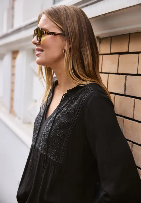 Vrouw zijwaarts tegen een muur, lang haar, draagt een zwarte blouse met kanten details.