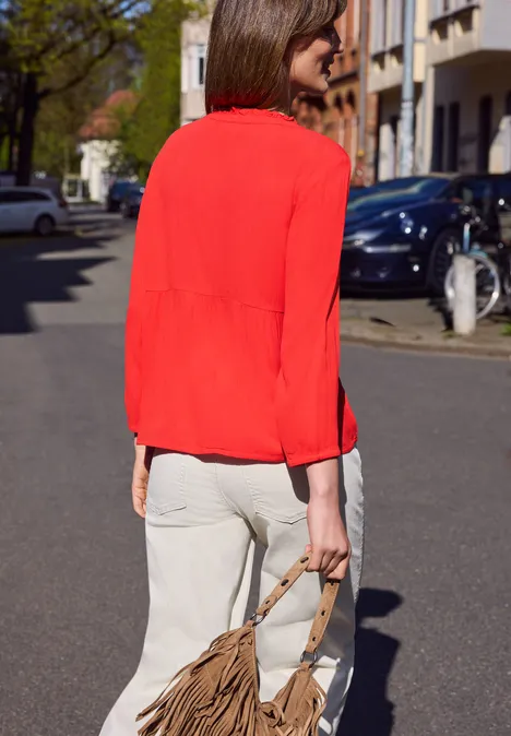 Een vrouw in een rode blouse en witte broek loopt op straat en draagt een bruine tas.
