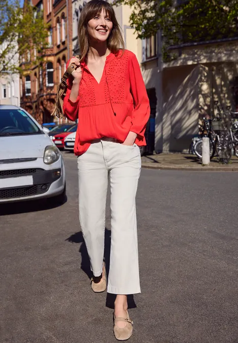 Een vrouw met een rode blouse en witte broek loopt over een zonnige straat.