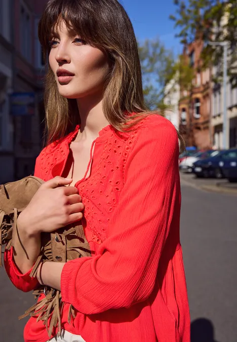 Een vrouw in een rood kanten bloesje houdt een bruine tas vast, staand op een zonnige straat.