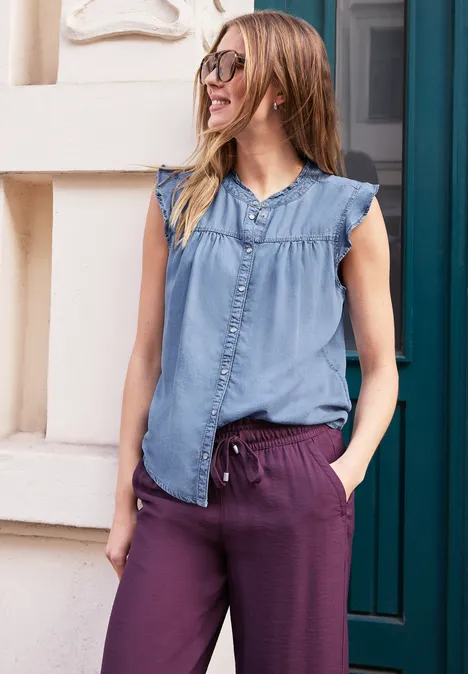 Frau in ärmelloser Jeansbluse und lila Hose lehnt entspannt an einer Hauswand.