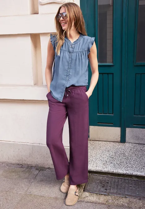 Frau in ärmelloser blauer Bluse und weiten violetten Hosen steht entspannt vor einem Haustürportal.