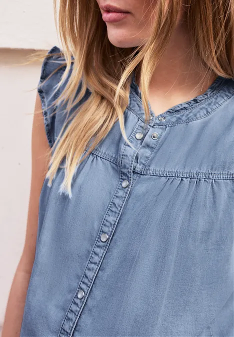 Frau in ärmellosem, hellblauem Denim-Top mit halblangen, blonden Haaren vor neutralem Hintergrund.