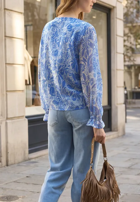 Vrouw van achteren in blauwe blouse met print, spijkerbroek en bruine franjes tas op straat.