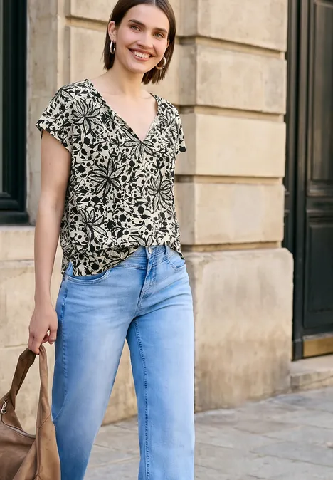 Vrouw in gemêleerd shirt, lichtblauwe spijkerbroek en tas, lopend langs een stenen muur.