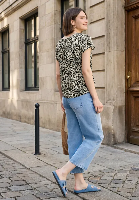 Vrouw in jeans, bloes met bloemen en blauwe sloffen loopt over een geplaveide straat langs een gebouw.