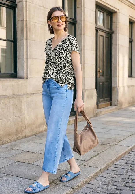 Vrouw in printshirt en jeans met sandalen en handtas loopt langs een gevel in de stad.