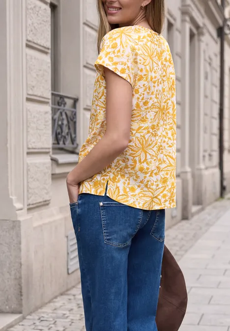 Junge Frau in gelbem Blumenoberteil und Jeans, die auf einer städtischen Straße steht und lächelt.