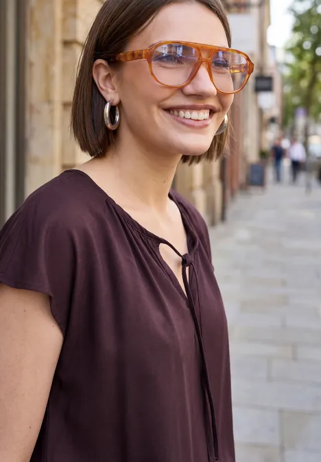 Frau in kastanienbraunem Oberteil mit silbernen Creolen steht auf gepflasterter Straße vor Häusern.