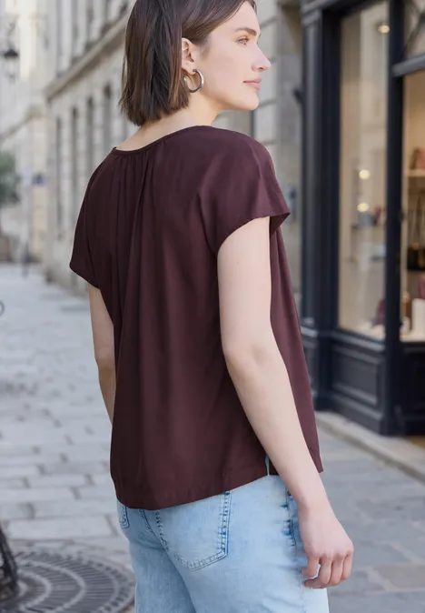 Frau in Burgunderfarbener Bluse und Jeans steht mit dem Rücken zur Kamera auf einer Kopfsteinpflasterstraße.