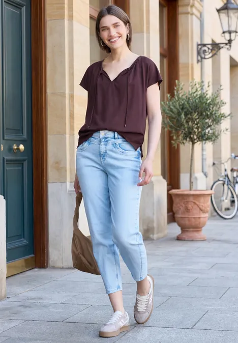 Eine Frau geht lässig vor einem Gebäude mit hellen Jeans, dunkler Bluse und Tasche.