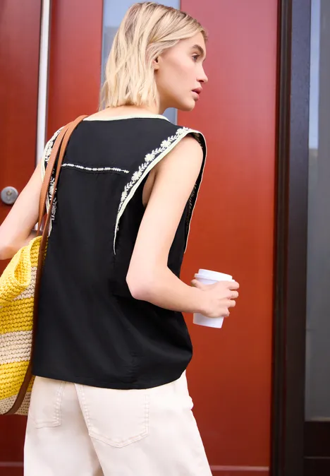 Blusentop mit Split Neck und Embroidery Black Frau in schwarz-weiß gesäumtem Top und weißer Hose trägt gelb-weiße Strandtasche und Kaffee vor roten Türen.
