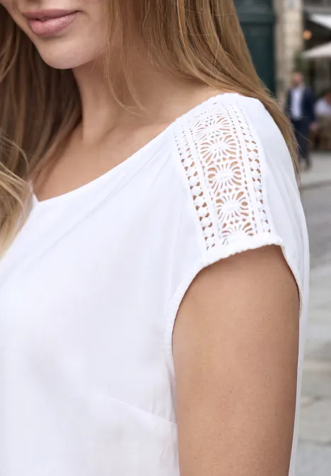 Une femme porte un top blanc à manches courtes avec des incrustations de dentelle élégantes sur les épaules.