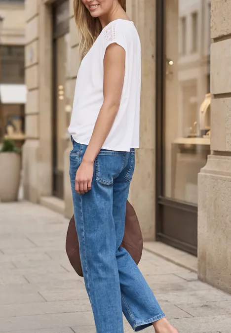 Une femme en haut blanc et jean bleu tient un chapeau derrière le dos sur un trottoir urbain.