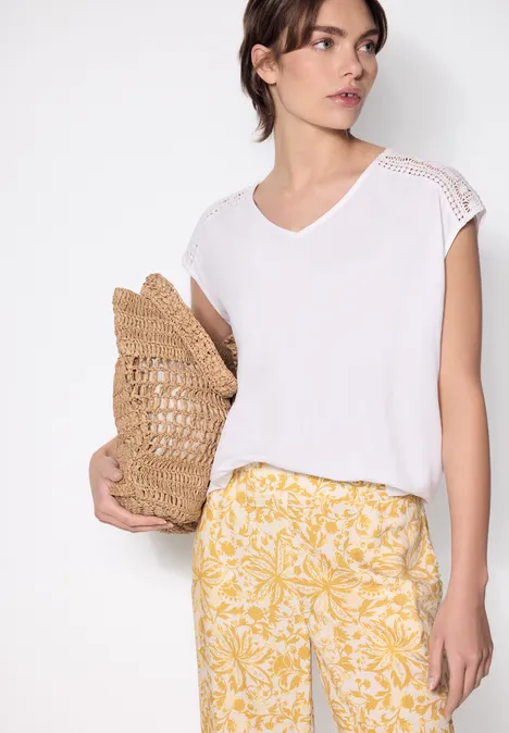 Femme porte un top blanc à col V avec dentelle, pantalon jaune à imprimé floral blanc et sac en paille.
