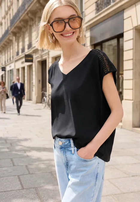 Eine Person in schwarzer Bluse und Jeans spaziert vor einer eleganten Steinfassade auf einer Stadtstraße.