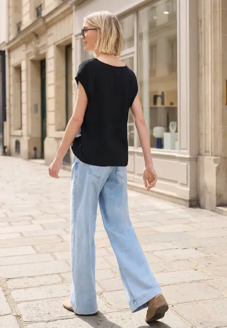 Frau mit Sonnenbrille, schwarzem T-Shirt und hellblauen Jeans läuft durch eine Stadtstraße.