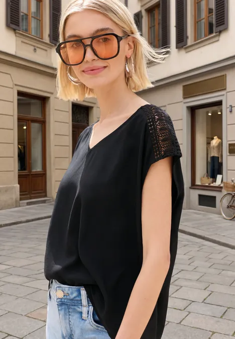 Eine Frau in schwarzem ärmellosem Top und Jeans steht in einer Kopfsteinpflastergasse vor Gebäuden.