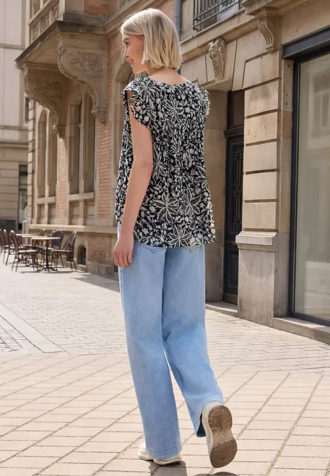 Frau mit schwarz-weiß gemustertem Shirt und hellblauen Jeans läuft auf gepflastertem Gehweg in urbaner Straße.