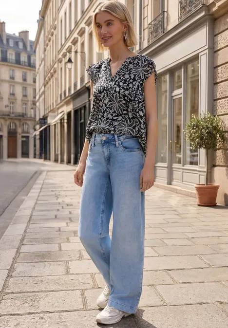 Eine Frau in gemusterter Bluse und weiten Jeans spaziert durch eine sonnige Altstadtgasse.