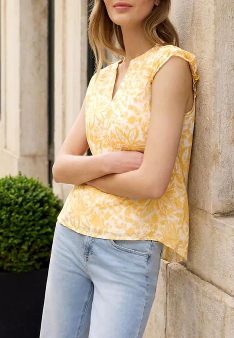 Frau lehnt seitlich an Steinwand, trägt gelbes Blumen-Top und blaue Jeans, Arme verschränkt, Pflanze im Hintergrund.