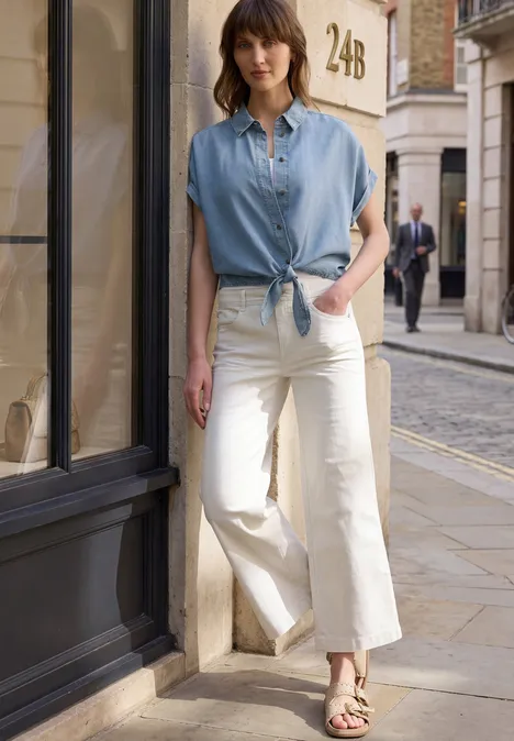 Frau in hellblauer Kurzarmbluse und weißen weiten Jeans steht auf der Straße.
