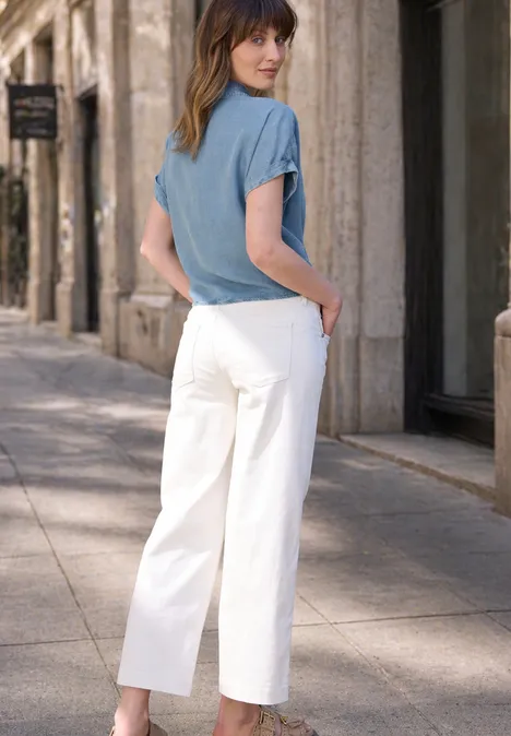 Frau in hellblauem T-Shirt und weißen, weit geschnittenen Hosen geht auf einem Bürgersteig an Gebäuden vorbei.