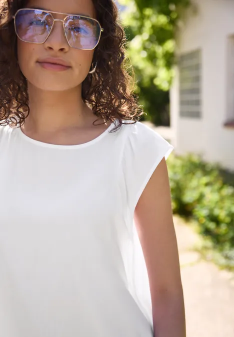 Dropped Shoulder Blusenshirt mit Rundhals White Eine Person mit lockigem Haar in weißem ärmellosen Oberteil im sonnigen Garten vor einem Haus.