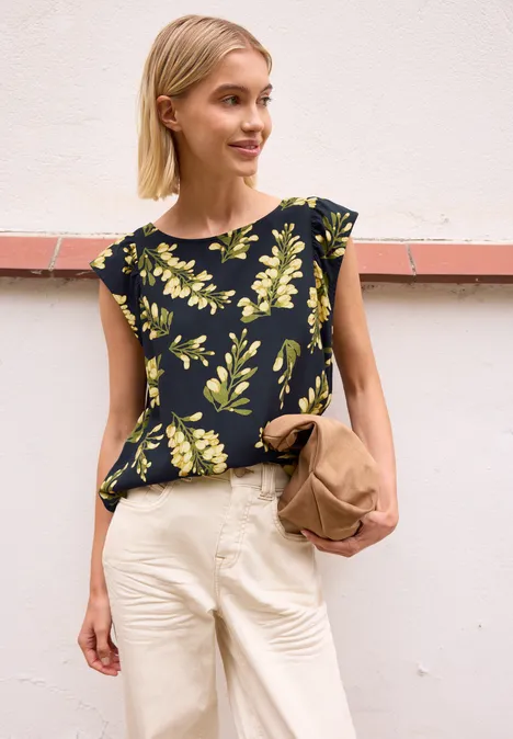 Vrouw in zwart bloesje met geel bladmotief, witte broek en bruine tas voor een lichte muur.