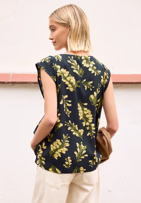 Vrouw van achteren in een donkerblauw bloesje met gele bloemen en lichte broek, houdt een tas vast
