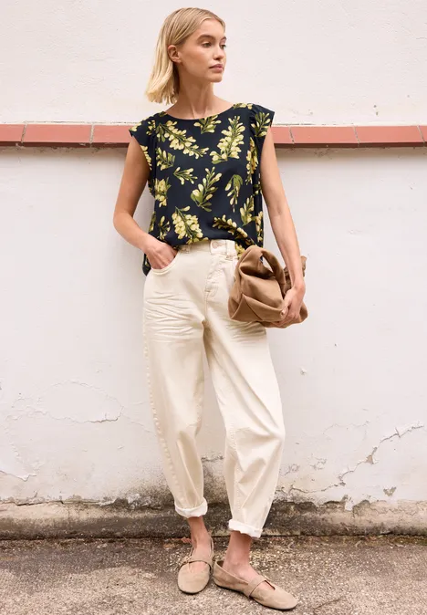 Vrouw voor lichte muur in losse zomerkleding: zwart bladprint top, wijde witte broek, sandalen en bruine tas