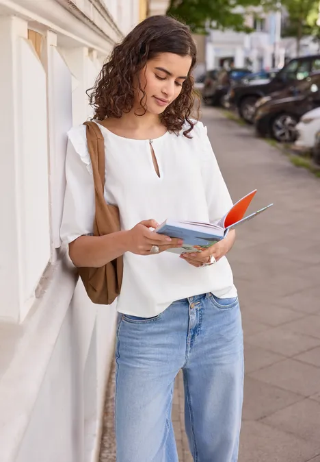 Kurzarm Bluse mit Volant-Details White Eine junge Frau lehnt an einer Hauswand und liest draußen in einem Buch.