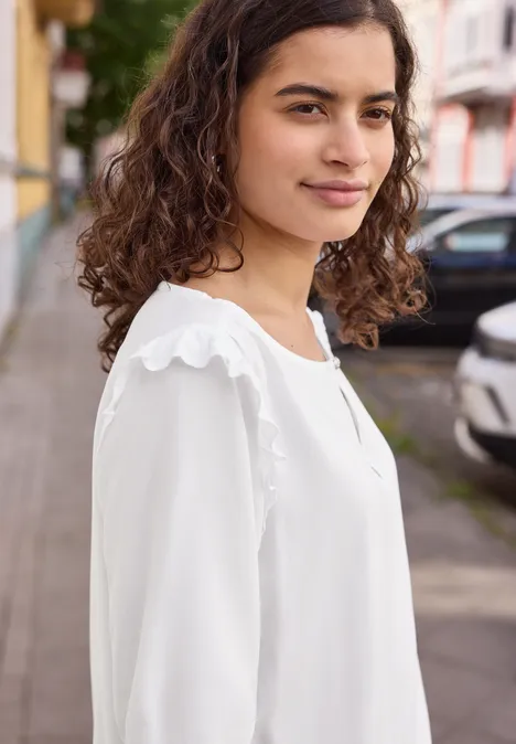 Kurzarm Bluse mit Volant-Details White Eine Frau mit lockigem Haar trägt eine weiße Bluse und steht auf einem Bürgersteig vor geparkten Autos.