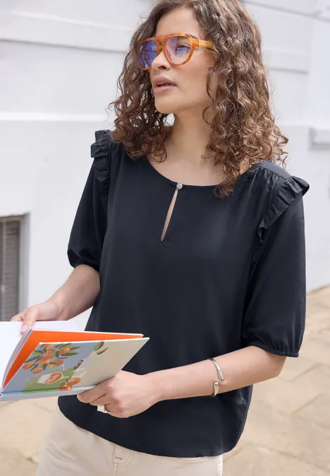 Frau mit lockigem Haar in schwarzer Bluse hält ein farbenfrohes Buch vor einem hellen Gebäude.