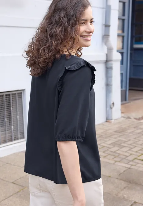 Kurzarm Bluse mit Volant-Details maritime blue Person mit lockigem Haar in schwarzer Bluse steht seitlich auf dem Bürgersteig vor Häuserfassade.