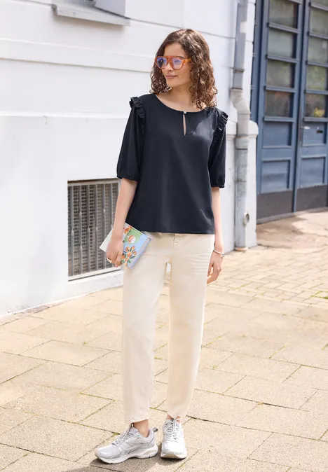 Kurzarm Bluse mit Volant-Details maritime blue Eine Frau trägt eine schwarze Bluse mit Rüschenärmeln, helle Hose, silberne Sneaker und hält eine bunte Tasche.